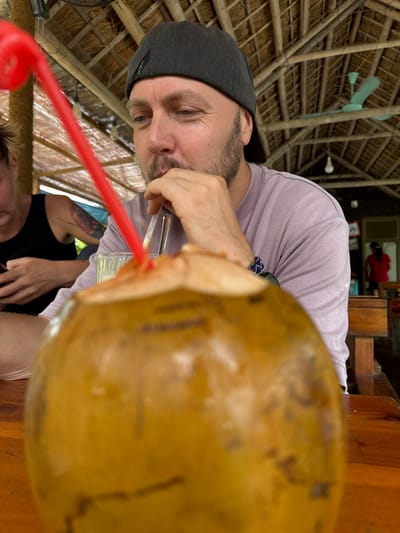 A man wearing a grey baseball cap and a light purple t-shirt is shown from the chest up, with his face in profile, looking down and to his left. He has a beard and is resting his chin on his hand, with a straw in his mouth. In the foreground, a whole coconut, cut open, is placed on a wooden table, obscuring the lower part of the image. A red straw is inserted into the coconut. To the left of the man, another person is visible from the shoulders up, wearing a black tank top and a tattoo on their arm, also looking down at a phone. The background shows a thatched roof structure with visible beams and a fan. In the distance, two figures are visible in a doorway. The setting appears to be an outdoor eatery or bar in Nam Hoa Lư Ward, Vietnam, suggested by the tropical architecture and the presence of a coconut drink.