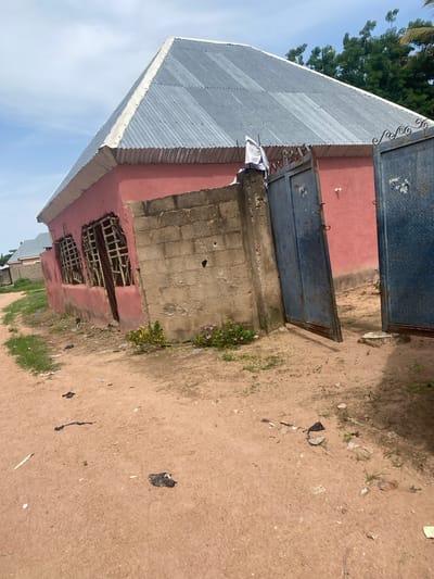 The image depicts a modest building with a corrugated metal roof and pink-painted walls. The building appears to be constructed from concrete blocks in some areas. There are two large, blue metal gates, one of which is open, revealing a further pink-walled structure behind it. Small pink flowers are growing near the building. The ground is sandy and somewhat littered with debris, including pieces of paper and what appears to be a black plastic bag. In the background, there are trees and a clear blue sky, suggesting it is daytime with fair weather. The scene has a rural or semi-rural feel, consistent with the provided location context of Jauro Yinu, Nigeria. No people are visible in the image. The overall impression is one of a simple, functional dwelling or establishment in a developing area.