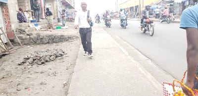 The image depicts an active urban street scene in Goma, Democratic Republic of the Congo.

In the immediate foreground on the right, the lower torso and legs of a person are visible, wearing blue shorts and holding two woven plastic bags, one red and white, the other yellow and white.

A paved sidewalk occupies the central foreground and midground, running parallel to a paved road on the right. A man in a white long-sleeved shirt, dark trousers, and white-soled sneakers walks towards the viewer on this sidewalk, carrying a dark backpack over his left shoulder.

To the left of the sidewalk, an unpaved dirt and gravel shoulder contains a pile of dark, irregular rocks or debris. Further left, elevated concrete steps lead to a storefront or building. Several individuals are present in this area; one woman sits on the steps in a patterned dress, while others stand nearby. One person, partially obscured, appears to be holding an infant. Blue and green plastic basins are visible on the ground near these individuals. Buildings with corrugated metal and painted walls line this side of the street.

The paved road on the right features multiple motorcycles in motion, typically with single riders, though some may have passengers. The motorcycles are various colors, and riders wear helmets or casual clothing. Beyond the road, additional urban structures, including multi-story buildings and market stalls, extend into the background. The scene suggests a busy commercial or residential thoroughfare.