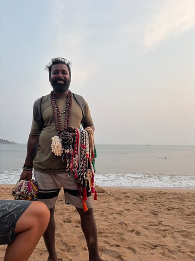 A dark-skinned adult male, barefoot, stands on a sandy beach in Gokarna, India, facing and smiling directly at the viewer. He has dark, curly hair and a full beard. He wears an olive green short-sleeved t-shirt, light grey shorts with dark horizontal stripes, and a dark backpack on his shoulders. Multiple strands of dark brown rudraksha beads are draped around his neck. He holds a significant quantity of necklaces, bracelets, and shell ornaments in both hands, including items made of colorful beads, shells, and tassels. The lower half of another person's legs, clad in grey patterned shorts, is visible in the extreme lower-left foreground. A light-colored dog lies partially obscured on the sand behind the man's legs. The background reveals a calm sea with small waves breaking on the sandy shore. A distinct rocky outcrop extends into the ocean on the far left. A dark, distant boat is visible on the horizon, and a solitary figure is in the water closer to the shore. The sky is hazy and light blue-grey.