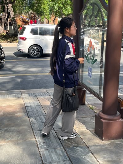 A woman with long dark hair, wearing glasses, a dark blue jacket with white and red stripes, gray pants, and black and white sneakers, is standing at a bus stop. She is looking into the bus stop's display case. She has a black purse slung over her shoulder. The bus stop is located on a paved area near a street with a white SUV parked alongside it. The location is likely in Changchun City, China, as evidenced by Chinese characters on the bus stop display. A decorative display of wheels is visible across the street.