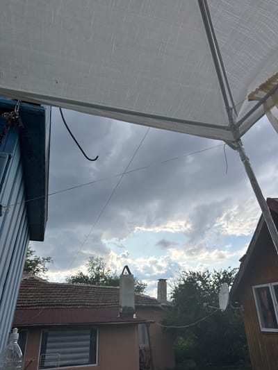 The image depicts a view from under a white tarp, looking towards a cloudy sky with patches of blue. In the foreground, a blue, corrugated metal structure is visible on the left, with a chain attached to it. A bent metal hook hangs from the structure. A thin wire stretches across the frame. In the middle ground, several houses with red tile roofs are visible. One house has two chimneys, and another has a satellite dish mounted on its side. Trees surround the houses. On the far right, a wooden house with a window is visible. The scene appears to be in a residential area in the city of Sredets, Bulgaria.