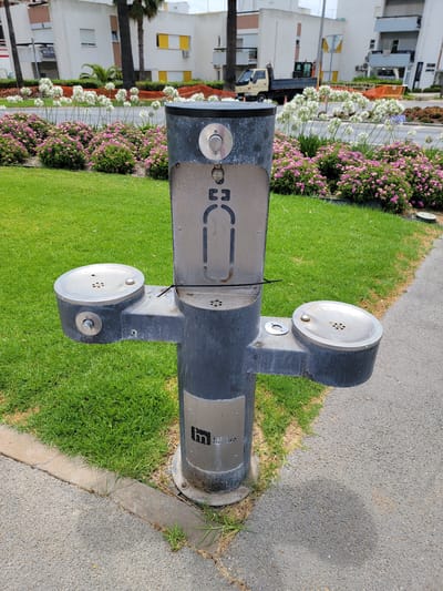 This is a medium shot, outdoor, eye-level, vertical photograph of a drinking fountain located in a public area. The fountain is dark gray with silver accents. It has a central horizontal pipe with two round, metallic dispensers on either side, each with a button and a grated opening. A metal plate with a stylized "L.F." logo is affixed to the right side of the central pipe. The fountain is situated next to a strip of lush green grass, which transitions to a paved walkway. In the background, there is a row of flowering bushes with pink and white blossoms, and behind them, several multi-story white buildings with balconies and windows. The sun is casting a shadow from the fountain onto the paved walkway, suggesting it is daytime. The overall setting appears to be a park or a public promenade in a warm climate. There are no people visible in the frame. The scene is bright and appears to be a sunny day, possibly in the late morning or early afternoon. The location context suggests the image was taken in Quarteira, Portugal.