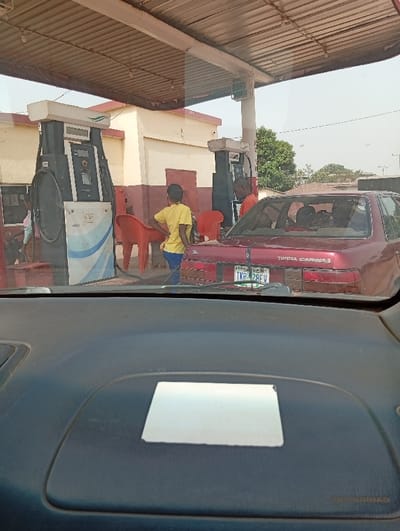The image depicts a petrol station in Peyer, Nigeria, viewed from inside a vehicle. The foreground shows a dark gray vehicle dashboard with a white rectangular object and an "SRS AIRBAG" label. In the midground, a red Toyota Carina II sedan, with the Nigerian license plate "TKP-128FV", is positioned at a fuel pump. A person wearing an orange shirt is visible at the rear of the red car, interacting with the fuel pump, suggesting fuel dispensing. To the left of the red car, another person in a yellow t-shirt and blue trousers stands with hands on hips, facing away from the camera. Two red plastic chairs are placed between the red car and a fuel pump. A large, dark gray fuel dispenser with a white and blue striped base and "MEDES" branding is prominent on the left. A third individual, partially obscured and possibly a child in light-colored clothing, is visible near this leftmost pump. The background features a cream-colored, single-story building with a reddish-brown base, and green foliage. A corrugated metal canopy covers the station area. The sky is clear.