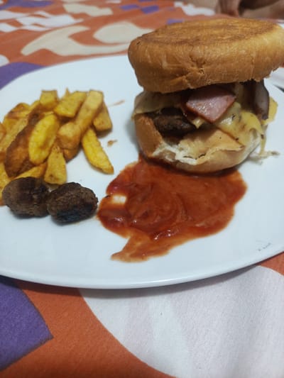 A plate of food sits on a patterned tablecloth. The food consists of a large burger with cheese and ham, a portion of french fries, two meatballs, and a pool of ketchup. The location is Guayaquil, Ecuador.