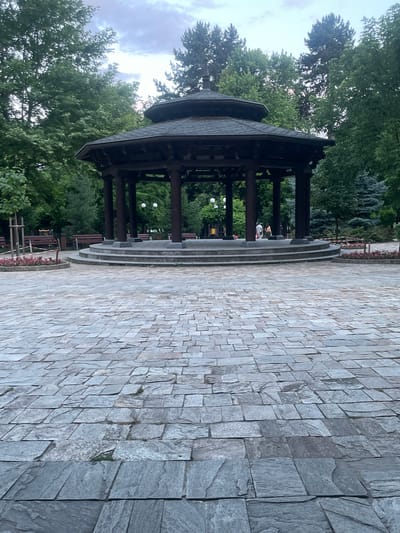 The image depicts a large, octagonal wooden gazebo with a dark, multi-tiered roof supported by thick pillars. The gazebo is situated on a raised, circular platform accessed by several steps. The platform and the area in front of it are paved with irregular, grey flagstones. Surrounding the gazebo are lush green trees of various species, with some evergreen trees also visible. In the background, there are park benches, lampposts, and a glimpse of playground equipment. Two individuals are visible walking near the gazebo. The scene appears to be set in a park, likely during twilight or a cloudy day, given the soft lighting and diffused sky. This is the central gazebo in the city of Gotse Delchev, Bulgaria, a common gathering place for residents.