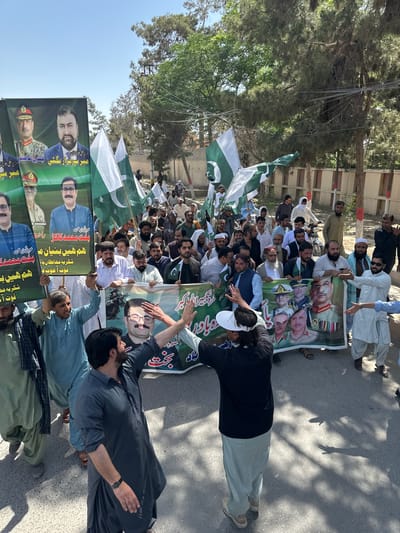 A large group of people, predominantly men, are gathered in what appears to be a street in Quetta City Tehsil, Pakistan. They are holding Pakistani flags and banners featuring portraits of various individuals, including military and political figures. The banners also have text in Urdu. The crowd seems to be participating in a rally or demonstration. Some people are wearing traditional Pakistani clothing, such as shalwar kameez, and others are in Western-style attire. The scene is bathed in sunlight, indicating it is daytime. The background includes trees and buildings, suggesting an urban setting.
