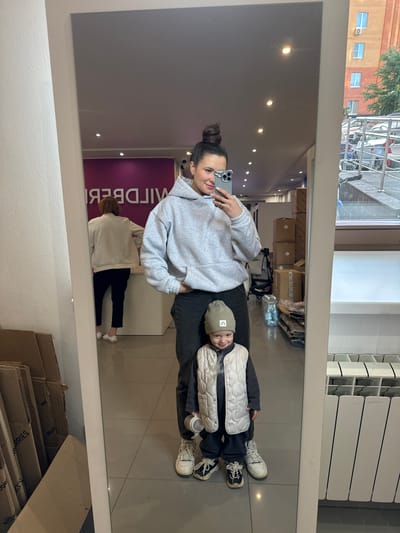 A photograph shows a woman and a child reflected in a mirror. The woman is holding a smartphone, presumably taking the picture. She has her hair in a bun and is wearing a light gray hooded sweatshirt and dark pants. The child is lying on the floor with their head near the woman’s hip, wearing a khaki hat, a padded vest, a dark gray shirt, dark pants, and sneakers. The background shows the interior of what appears to be a store, with boxes stacked in the back and a woman reflected in the mirror wearing beige top and black pants. Outside, there is a building with windows and trees, and a street with parked vehicles. A sign with the store's name "WILDBERRIES" is visible in mirror. Based on this information, the location is likely in a Wildberries store in the city of Odintsovo, Russia.