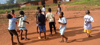 The photograph depicts a group of approximately ten children in what appears to be a dirt road or open area in Ngalula, Democratic Republic of the Congo. Several buildings, possibly residences, are visible in the background. The children are of African descent and are dressed in various casual clothing, including t-shirts, shorts, and sandals. One child is wearing a white soccer jersey with the name "BENZEM" and the number "9" on the back. The children are interacting with each other, some touching or gesturing. The ground is reddish-brown dirt. The environment suggests a rural or semi-rural setting. There is some vegetation visible in the background near the buildings.