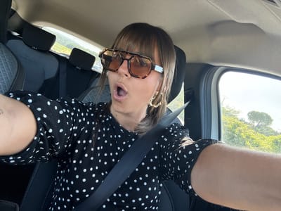 A woman with bangs, wearing large, brown-tinted, tortoiseshell sunglasses and a black dress with white polka dots, is taking a selfie in the driver's seat of a car. She has an open-mouthed expression and is wearing large, gold hoop earrings. Her left arm is extended, and her right arm is out of view. The car's interior, including seats and a seatbelt, is visible. The exterior view through the window shows a sunlit, tree-lined landscape, likely in the vicinity of Le Muy, France, given the geographical context.