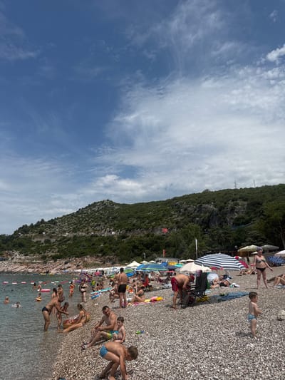 This image depicts a lively beach scene in Kamenovo, Montenegro. The beach is covered in small, grey pebbles, and the water is clear and calm. Numerous people are present, enjoying the sunny weather.
In the foreground, several individuals are on the beach, some sitting under umbrellas, others wading in the shallow water. A man in red shorts is tending to a beach chair, while a young child in blue swim trunks plays on the pebbles. To the right, a woman in a black bikini stands near an umbrella.
Further back, the beach is more densely populated. People are spread out on towels and beach chairs, with many in the water, swimming or playing. Inflatable toys, including a large donut-shaped float, are visible. Beach umbrellas of various colors and designs provide shade.
The background features a rocky, tree-covered mountainside that slopes down towards the sea. The vegetation on the hillside is dense and green. A road can be seen winding along the mountainside, with some small buildings and structures visible in the distance. The sky is blue with scattered white clouds.