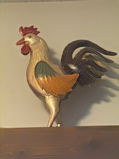A metallic rooster ornament, predominantly gold, stands on a dark brown wooden shelf against a light grey wall. The rooster has a red comb and wattle, a red beak, and its wings are detailed with green and orange feathers. Its tail is composed of dark, curved metallic feathers, each dotted with white. The ornament casts a shadow on the wall behind it. The image does not provide any information about the location within the Town of Cochrane, Canada.