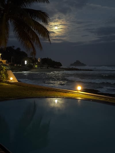 This media file depicts a nighttime scene at a beachside location, likely in Ahangama, Sri Lanka, characterized by a full moon partially obscured by clouds and illuminated by artificial lights. The foreground features the silhouette of a palm tree with fronds swaying, suggesting a gentle breeze.
The scene transitions to a sandy beach with rough waves breaking on the shore, creating a dynamic visual texture. Beyond the beach, a collection of trees and some structures are visible, including streetlights that cast pools of warm light. The presence of a retaining wall topped with grass runs along the right side of the image, bordering a large body of water, possibly a swimming pool or calm inlet, reflecting the ambient light.
The overall mood is serene and slightly mysterious, amplified by the interplay of natural moonlight and man-made illumination. The time of day is clearly night, and the weather appears calm, despite the suggestion of wind from the palm fronds. The location cues, such as the palm trees and tropical foliage, along with the beach and the specific style of the retaining wall, point towards a coastal resort or residential area in a warm climate. No text is visible in the image.