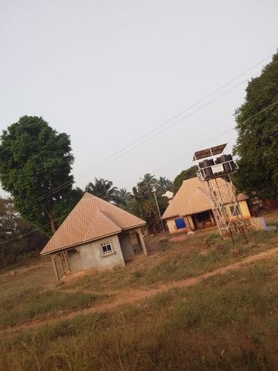 This image depicts a semi-rural landscape in Ankpa, Nigeria, under an overcast, light grey sky. The ground consists of reddish-brown soil with scattered patches of dry and green low-lying vegetation.
Two single-story structures with light brown corrugated metal hip roofs are visible. The building on the left features unpainted grey concrete block walls, a single rectangular window with white framing, and an open front porch supported by pillars. To its right, another building with a similar roof appears; a blue tarp or fabric covers a portion of its exterior, possibly an entrance or window.
Between these two buildings, a tall metal lattice tower stands, supporting two black cylindrical water tanks and a framework of several dark rectangular solar panels beneath a light-colored canopy, indicative of a solar-powered water system. A green sign with white text is affixed to the tower's support structure. A tall street light pole with a luminaire is positioned near the second building.
Overhead, three thin electrical wires run horizontally across the frame. Large, leafy green trees, including discernible palm trees, are present on the left and right edges, as well as in the background. No individuals are present in the scene.