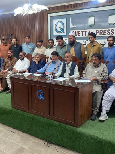 A group of men are seated at a large brown desk in what appears to be a press conference setting. The background features a sign that reads "QUETTA PRESS." The setting suggests this event is taking place in Quetta, Pakistan. The men are dressed in traditional Pakistani clothing, and some are holding papers, possibly reading from prepared statements. The overall scene conveys a formal gathering, likely for an official announcement or press briefing.