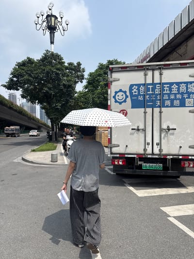In a city scene in Tianhe District, China, a person walks away from the camera under a white umbrella with black polka dots, holding a folded piece of white paper in their left hand. They are wearing a gray t-shirt and black and white striped wide-leg pants. The person is walking on a crosswalk in front of a large white truck. The truck has blue Chinese characters and a blue logo on its side. The license plate of the truck is green and reads "A B41036". Behind the truck, a white building with a corrugated roof is visible. To the left of the truck, trees line the sidewalk and street. In the background, a highway overpass with cars can be seen, and tall buildings are visible in the distance. A traditional lamppost with multiple lights stands above the trees. The sky is clear and blue.