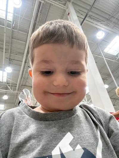 A close-up, low-angle, selfie-style shot shows the face of a young Caucasian boy with light brown hair. He is wearing a gray crew-neck sweatshirt with a white and navy blue graphic design on the front. The boy is looking down and smiling slightly. The background appears to be an indoor public space with a high ceiling, exposed industrial-style beams, and fluorescent lighting. A large white pillar is visible on the right side of the frame. A small portion of another person's arm, wearing a red sleeve, is visible in the far right background. The image is taken from an upward perspective, with the boy's chin and shoulders prominently in the foreground.