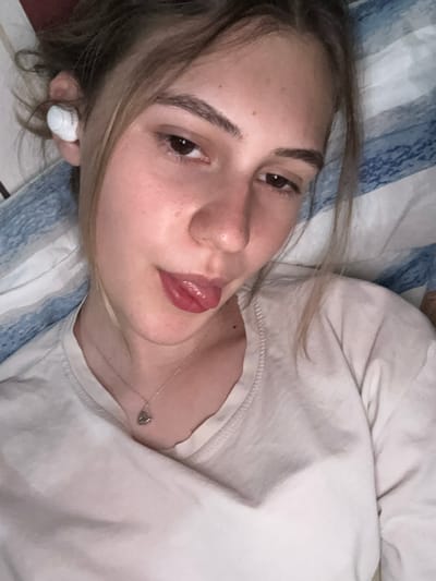 The image shows a close-up, angled selfie of a young woman lying down, with her head slightly tilted and her tongue sticking out. She has light brown hair pulled up loosely, and is wearing white earbuds in her left ear. She is wearing a light-colored t-shirt and a delicate silver necklace with a heart-shaped pendant. The background appears to be a bed with blue and white striped bedding. The overall lighting is soft and appears to be artificial, casting a warm glow on her face. There is no discernible location in the city of Panagyurishte, Bulgaria, or any other specific geographical markers in the image.