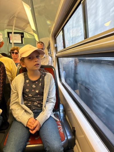 A child with a pink baseball cap sits on a train seat. They wear a grey sweater and jeans over a dark grey Mickey Mouse shirt. An older man and woman are in the background. Light enters through the train’s overhead windows. The child looks directly into the camera. The interior of the train is visible. The scene appears to be a candid shot.