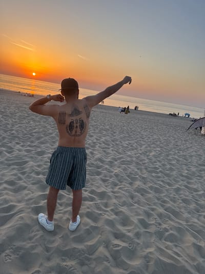 A man stands on a sandy beach at sunset, his back to the camera. He is wearing a dark baseball cap, striped shorts, and white sneakers. His back is covered in intricate black tattoos, including two prominent tiger designs at the top, surrounded by geometric patterns and script.
The sun is low on the horizon, casting a warm orange glow across the sky and reflecting on the calm ocean. The beach is composed of rippled sand, with footprints visible in places. In the distance, the ocean stretches out, and a faint line of people and beach equipment can be seen along the shoreline. The sky transitions from a pale blue at the top to soft orange and yellow hues near the horizon.
The overall mood is serene and peaceful, with the natural beauty of the sunset creating a tranquil atmosphere. The man appears to be enjoying the moment, with one arm raised slightly. The location is identified as The Hague, Netherlands. No visible text is present in the image.