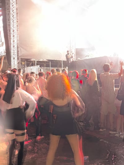 The image captures a vibrant outdoor music festival or concert scene, possibly at a location within the Palilula Urban Municipality of Serbia. A large, brightly lit stage or performance area is visible in the background, with a massive screen or projection surface dominating the upper portion of the frame. The lighting is intense, creating a hazy and ethereal atmosphere with strong backlighting that obscures finer details and silhouettes many of the figures.
In the foreground and midground, a crowd of concertgoers is densely packed, engaged in the lively atmosphere. Many individuals appear to be dancing or interacting, with a sense of movement conveyed through motion blur. The attire of the attendees is varied, ranging from casual festival wear to more stylized outfits. Several individuals are clearly visible, including a woman in the center with curly, reddish-brown hair wearing a black dress and another person in the foreground on the left wearing a white top and black skirt with stockings. The ground appears to be wet or reflective, possibly due to water or spilled drinks, with some metallic grating visible in the lower portion of the image.
The overall impression is one of energetic celebration and a shared experience at a large-scale event. The presence of stage equipment, lighting rigs, and a large audience strongly suggests a music performance or festival setting.