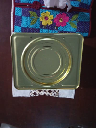 A metallic, square tin rests atop a white cloth with a repeating pattern of brown and white ovals. A quilted, blue and purple patterned fabric with embroidered yellow and pink flowers is partially visible in the upper background. The fabric appears to be used as a tissue holder. The entire scene is set against a dark brown surface.