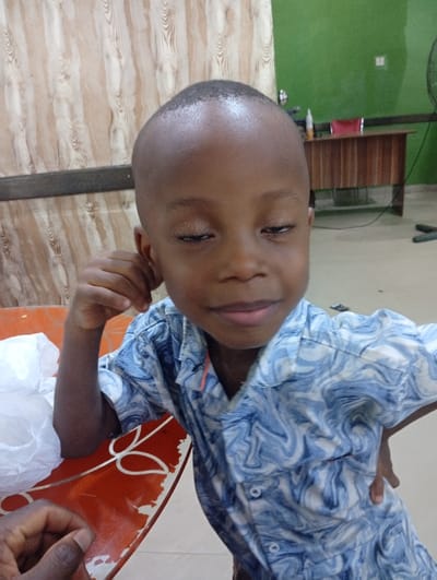 A young boy with a shaved head and closed eyes sits at a table, wearing a blue and white patterned shirt. He rests his head on his right hand. Part of another person's hand appears in the lower left corner of the frame. A red table with white accents holds a crumpled white plastic bag. Behind the boy is a wall, half covered in brown wood paneling and half in green paint. In the background, there is a wooden desk, with a few objects on top.