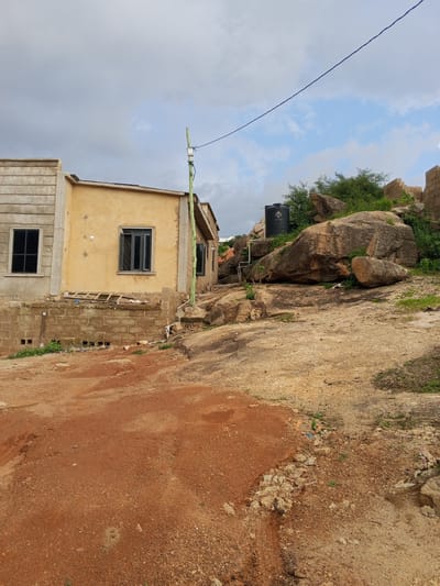 The image captures an outdoor scene in Kabong, Nigeria, featuring a partially constructed building and a rocky, uneven terrain. The building on the left appears to be a single-story structure with a yellow-painted facade and a section with textured concrete walls. It has dark-framed windows. Adjacent to it, to the right, is another section of the building, also under construction, with visible brickwork.
The foreground is dominated by a reddish-brown, dusty ground, likely soil or gravel, with some scattered debris. The middle ground is characterized by a large, weathered rock formation with various sizes of boulders and smaller stones, interspersed with green vegetation. A tall, black water tank is positioned on top of the rocks. A green pole with electrical equipment is visible near the building, and a blue wire stretches across the upper part of the frame. The background consists of a cloudy sky with patches of blue. The overall impression is of an undeveloped or developing area.