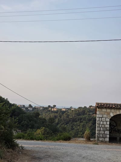 This image shows a view from a dirt road in Vence, France. In the foreground, the road is rough and uneven. To the right, there is a small stone structure with a tiled roof and an arched opening, which has graffiti on the inside wall. Behind the structure, a dense green forest covers a hillside. In the distance, several houses are visible nestled on the slopes, and the sea can be seen beyond them. Overhead, power lines stretch across the sky. The sky is pale and overcast. There are no people or animals visible in the scene.