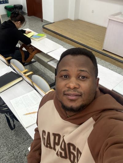 The image depicts an indoor classroom setting, presumably located in Daxing District, China. In the foreground, a male individual of African ethnicity is taking a selfie, looking directly at the camera with a slight smile. He has short, dark hair and a beard, and is wearing a two-tone hoodie, brown on the sleeves and hood, and beige on the torso with "GAP 1969" embroidered in brown letters.

In the mid-ground, a female individual with dark hair tied back is seated at a student desk, facing away from the camera. She is wearing a black long-sleeved top and dark trousers. Her attention is directed downwards towards an open yellow book and a green book on her desk. A brown shoulder bag is visible hanging from the side of her desk.

The classroom features multiple rows of light-colored student desks with attached chairs. On the desk directly in front of the male individual, an open notebook with handwritten text and a pencil is visible, alongside a closed black book. A black bag/backpack is positioned under this desk.

The background shows a raised wooden platform or stage area. To the left of the platform, against a white wall, two trash bins, one green and one black, are visible. Above these bins, a portion of a whiteboard or wall-mounted panel with numbers "58," "59," and "60" is discernable. The walls are predominantly white, and the flooring in the main seating area appears to be a light-colored, possibly patterned, surface.