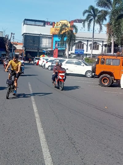The image shows a street scene in Pati, Indonesia, taken from the perspective of a person standing in the middle of the road.  A man is riding a black bicycle on the left side of the image, heading toward the viewer. To the right of the bicycle, a person is riding a red motorcycle toward the viewer.  Behind the cyclist, there is a parked row of cars, including a white sedan, a black SUV, and a large orange truck with a large tire.  In the background, there are buildings, including a large building with a blue and yellow sign on it.  The sky is clear and blue.  The image was taken at coordinates -6.754 latitude and 111.039 longitude.
