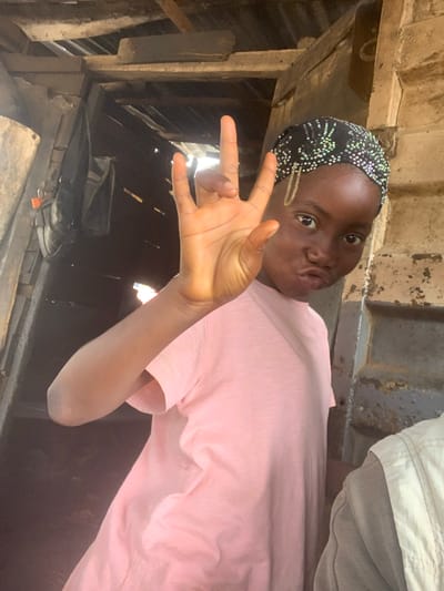 The media file is a portrait of a young Black individual, likely female, wearing a pink t-shirt and a black headscarf adorned with small, shiny beads. The person is positioned in a slightly reclined, informal pose, making a "peace" sign with their right hand. Their lips are pursed, suggesting a playful or coquettish expression. A golden hair clip is visible on the left side of their forehead, adding a subtle decorative detail. The background is rustic and appears to be an outdoor or semi-outdoor setting. There are wooden structures and possibly corrugated metal elements, indicating a possibly improvised or informal shelter. Beneath the subject, what appear to be shoes are visible in a dark, cluttered area. The lighting suggests it might be daytime, with some natural light filtering into the scene. The overall impression is of a casual, candid moment captured in what seems to be a less developed environment. The location context of Shagamu, Nigeria, suggests a West African setting.
