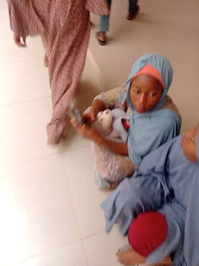 The scene depicts an indoor environment with a light-colored tiled floor in the city of Kwazailewa, Nigeria. A young female, appearing to be a child or teenager, is seated in the central foreground, wearing a patterned garment, a light blue hijab, and an orange head covering. She is observed holding two objects: a small, light-colored toy resembling a doll with a brown head and grey body in one hand, and a dark, rectangular object, possibly a mobile phone, in her other hand. Her gaze is directed downwards towards these items. To her immediate right, another individual is partially visible, seated, clad in a blue garment with a red item positioned on their lap; only their bare feet are discernible in the lower right. In the background, two additional individuals are in motion. The lower body of one person, dressed in a long, pleated, reddish-brown garment, walks from left to right. Further back, the feet, wearing dark sandals, and lower legs, clad in light blue trousers, of a fourth person are partially visible.