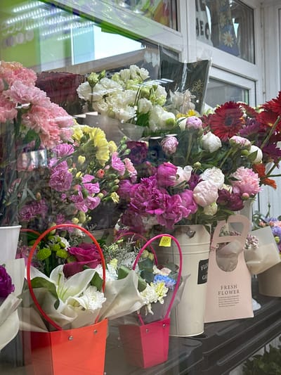 This media file displays a close-up, angled view of a vibrant assortment of flowers, likely in a shop or market setting. The scene is dominated by a variety of blooms in shades of pink, white, red, purple, and yellow, including what appear to be gerbera daisies, carnations, and possibly peonies and lilies. The flowers are arranged in bouquets and are partially contained within decorative packaging, such as a light beige metal planter and brightly colored pink and orange bags.

Several items of packaging are visible with text. One pink bag has the words "FRESH FLOWER" printed in a stylish font, along with accompanying descriptive text. A beige metal planter has "FLO" and "GAR" visible, likely part of "FLOWER GARDEN." There is also a small yellow tag attached to a pink ribbon, with some handwritten text that is illegible.

The overall impression is one of abundance and color. The lighting suggests it could be daytime, possibly indoors with reflections from windows or display cases. The reflections also reveal glimpses of a window frame and possibly other store merchandise, suggesting an interior retail environment. No people are visible in the frame. The setting appears to be a flower shop or stall.