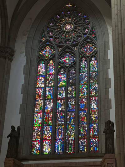The image shows a large, ornate stained-glass window inside a building, likely a church. The window is divided into sections: a circular rose window on the left and several rectangular panels on the right. The panels depict various scenes with figures and objects. The window is framed by gray stone architecture, including moldings and decorative sculptures. Two stone gargoyles or angel sculptures are positioned on the right side of the frame. The location is assumed to be in the city of São Paulo, Brazil.