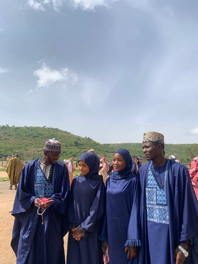 The image shows a group of people in Naraguta Hausawa, Nigeria. Four people are in focus, all wearing blue garments. The two men are in traditional attire with detailed embroidery. The two women wear hijabs. The background shows a partly cloudy sky and a green hillside. The scene is captured outdoors, likely during the day.