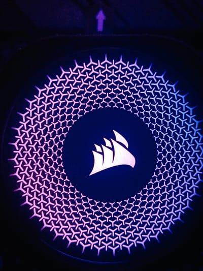 This is a close-up, eye-level shot of a black circular object, likely a computer fan grill, illuminated by purple and blue LEDs. The light emanates from a gradient, starting with a vibrant pink-purple on the left side and transitioning to a cool blue on the right. The grill itself is composed of a repeating geometric pattern resembling stylized "Y" shapes, forming concentric circles.
Centered within the grill is a bright white logo. It depicts a stylized silhouette of a sailing ship with three sails, set against a dark background. An upward-pointing purple arrow is visible in the upper portion of the image, above and slightly to the left of the circular grill.
The lighting creates a high-contrast aesthetic, with deep shadows emphasizing the illuminated patterns and logo. The overall mood is modern and tech-oriented, suggesting the object is part of a computer or gaming peripheral. There is no discernible information about the time of day, weather, or specific location within Ashkelon, Israel, beyond the implied context of a technological device. No text is visible in the image.