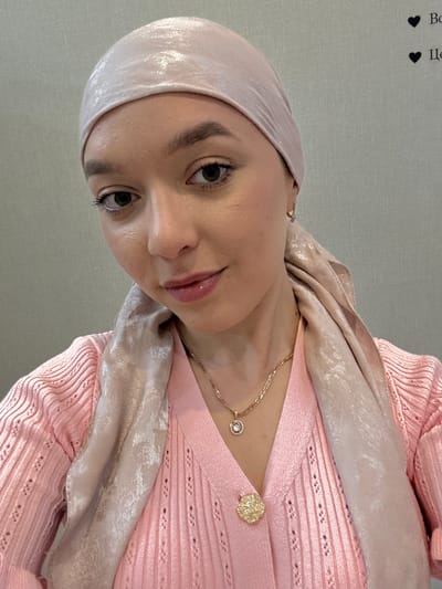 A woman is centered in the image wearing a pink headscarf, a gold necklace with a diamond pendant, and a pink knit sweater with a gold floral brooch. The background is a solid light gray color. The location is not discernible as Урмаево, Russia.