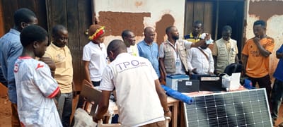 The image depicts a group of eleven men and one woman standing outside a building with rough, textured walls and wooden doors. The scene appears to be in Ngalula, Democratic Republic of the Congo.
The individuals are gathered around a table displaying various items. Prominently featured are solar panels, a large battery, two laptops, and what appears to be a blue cylindrical object wrapped in plastic. Several people are looking at the items on the table, and one man in a tan vest is gesturing towards them.
The attire of the individuals varies. Some are wearing casual clothing like t-shirts and button-down shirts, while others are in vests and what seem to be work uniforms. The woman in the foreground is wearing a distinctive knitted hat with red, yellow, and green accents.
The overall environment suggests an outdoor setting, possibly a community or training area, given the presence of what seems to be new equipment. The lighting indicates it is daytime.