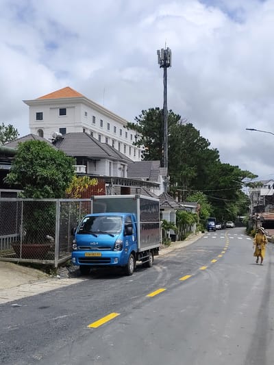 The image depicts a street scene in Lâm Viên - Đà Lạt, Vietnam, under a cloudy sky. In the foreground, a bright blue Kia truck is parked on the left side of the road, partially obscured by a chain-link fence. The truck has a silver cargo box. The road itself is asphalt with yellow lane markings.
Further down the street, several cars are visible. A woman wearing a yellow dress and a hat is walking away from the camera on the right side of the road. To the left of the road, there are houses with tiled roofs and lush green trees. In the background, a tall cell tower stands prominently, and a multi-story white building with a terracotta-tiled roof is visible. The overall impression is that of a residential street with urban infrastructure.