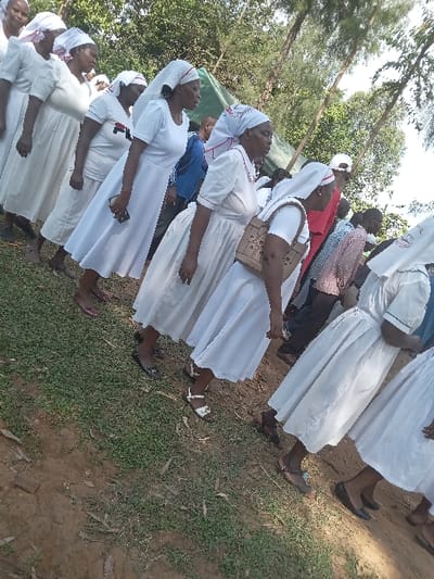 The image shows a group of women in white dresses and head coverings standing in a grassy area in Malava, Kenya. The women are arranged in a line with their backs to the camera, and the nearest woman's white head covering has a red trim. The women are standing on a dirt path surrounded by green grass, and trees are visible in the background. The women are likely attending a religious or community event.  The woman in the center of the image holds a brown handbag. There is a large white tent visible in the background to the right.  A partial view of another woman wearing a red, white, and blue dress is also visible in the background.  The coordinates for this location are 0.517 latitude and 34.821 longitude.