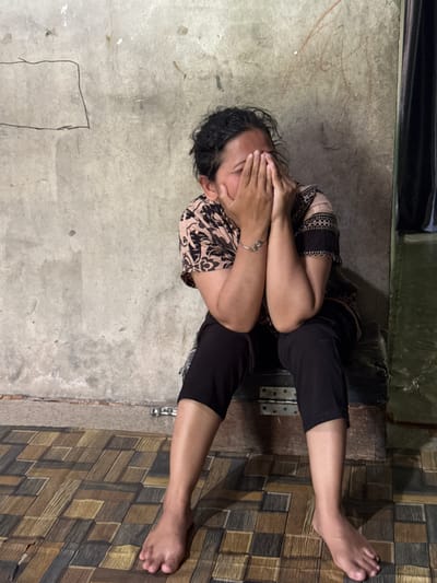 A woman with dark, curly hair sits on a dark-colored object, possibly a chest, against a rough, concrete wall. She is wearing a light-colored, patterned short-sleeved shirt and black pants. Her hands are covering her face, obscuring her eyes and nose, suggesting she might be crying or upset. A silver bracelet adorns her left wrist. Her bare feet are visible, resting on a floor with a tiled pattern of brown, beige, and gray rectangles. The environment appears to be indoors, with a dark curtain visible to the right. The wall has some markings, including a drawn rectangle and what appear to be faint red scribbles. The lighting is dim, casting shadows. The image is taken from a medium distance, capturing the woman from the knees up. The location is identified as Babahjurong, Indonesia.