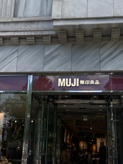 The image shows the exterior of a MUJI store, indicated by the prominent "MUJI" logo with Japanese characters above the entrance. The building's facade is made of marble, with decorative architectural elements above the store's awning. The store features a large glass entrance with reflective surfaces, allowing a partial view into the interior where clothing racks and displays are visible. Several people can be seen inside the store, some browsing merchandise and others interacting. The location is identified as MUJI store at Friedrichstraße 112, 10117 Berlin, Germany.