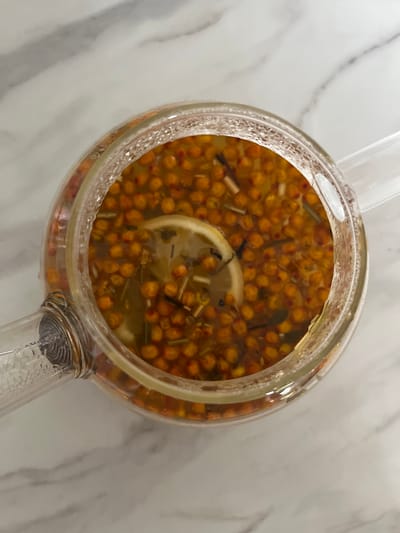 A close-up, overhead shot captures a glass teapot filled with a golden-orange liquid, teeming with small, round berries and what appear to be sprigs of herbs. A thin slice of lemon floats on the surface of the beverage. The teapot rests on a light-colored, marbled surface. The image does not contain enough information to determine the location in Novosibirsk, Russia, or any specific details about people, actions, or interactions.