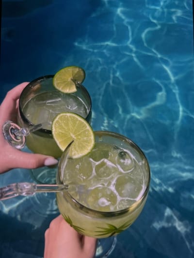 Two glasses of iced green liquid, likely cocktails with lime garnishes, are held by hands over a body of water with refracted light patterns. The glasses appear to be margarita glasses with stems. The environment suggests an outdoor setting, possibly a swimming pool. The location in Vidin, Bulgaria is not discernable from the image.