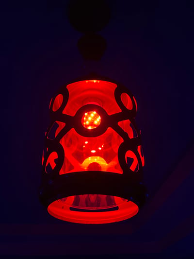 An illuminated, red, cylindrical object is suspended against a dark blue background. The object appears to be a lamp or lantern, with a decorative outer casing featuring circular and curved cutouts. Through these openings, a bright red light emanates from within, revealing a cluster of smaller, circular lights at the center. The light creates a warm, glowing effect, with reflections visible on the inner surface of the object. The overall impression is one of ambient illumination, possibly in a dimly lit indoor setting. The time of day and specific location are not clearly indicated, but the dark background suggests it is nighttime or that the environment is intentionally darkened. There is no visible text in the image. The object is positioned horizontally, with its left side slightly obscured by a dark shadow.