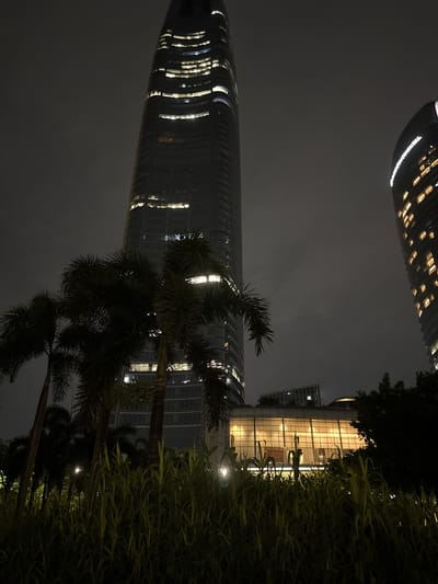 This media file is a nighttime photograph of a city skyline, with a focus on modern architecture and lush vegetation. The primary subjects are two towering skyscrapers, one significantly taller and more curved than the other. The taller building, on the left, has a sleek, dark facade illuminated by rows of warm yellow lights from within its many windows. Its reflective surface creates a sense of depth and grandeur. The other building, on the right, is wider and has a more geometric, glass-fronted design, also brightly lit from within, showcasing multiple floors and architectural details.
In the foreground, the scene is softened by the presence of tropical foliage, including palm trees and dense, grassy plants. These elements are silhouetted against the illuminated buildings and the dark, overcast sky, adding a natural contrast to the urban landscape. The overall effect is one of a vibrant, modern city at night, possibly a business or entertainment district.
The time of day is clearly night, indicated by the artificial lighting of the buildings and the dark sky. The weather appears to be cloudy or hazy, as the sky is uniformly grey and lacks any stars or clear celestial features. This atmospheric condition might contribute to a slightly diffused, moody lighting effect.
There is visible text on the upper left portion of the taller skyscraper that reads "INTERCONTINENTAL" in white, illuminated lettering. This suggests the building is a hotel. The location context provided (Haizhu District, China) further grounds the image in a specific urban environment. The combination of impressive architecture, vibrant lights, and the surrounding greenery creates a visually striking image that evokes a sense of sophisticated urban living and perhaps a touch of serene, tropical ambiance within the city.