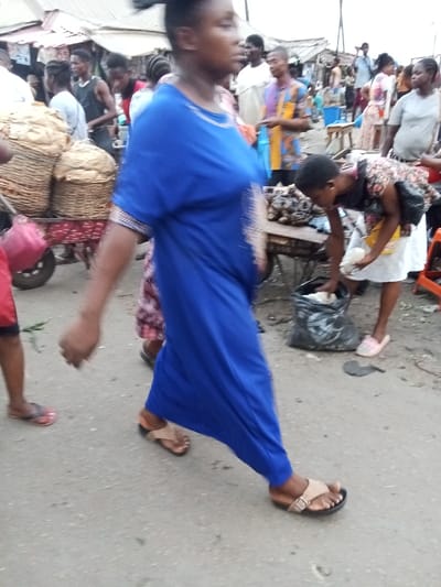 The image portrays an outdoor market scene on a paved surface in Owerri, Nigeria. In the immediate foreground, a dark-skinned individual, likely female, wearing a royal blue long dress with light embroidery and tan buckled sandals, walks towards the right. Her head is turned slightly to the right. To the right of this central figure, a young dark-skinned female, dressed in a floral top, white skirt, and pink flat shoes, bends forward. She is reaching into a black plastic bag positioned on the ground with her right hand, while holding a small white object in her left hand. A makeshift wooden table directly in front of her displays numerous dark, irregularly shaped items.

In the far left foreground, the lower torso and legs of a person wearing red shorts and red flip-flops are visible, holding green leafy material. Further into the mid-ground on the left, a pushcart laden with several large woven baskets, which contain bundled goods, is visible. The background is populated by numerous dark-skinned individuals, including a male wearing a multi-colored patterned shirt, standing amidst various market stalls topped with corrugated metal roofs. The scene conveys a bustling commercial environment.