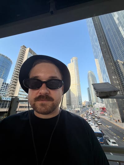 A man is taking a selfie on a street in Ramat Gan, Israel. He is wearing a black bucket hat, black oval sunglasses, and a black t-shirt. He has a mustache and a beard. In the background, there are tall modern buildings and a busy street with cars and buses. The sky is clear and blue.