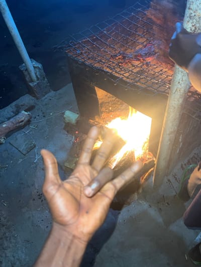 The image displays a nighttime outdoor scene in Birnin Kebbi, Nigeria, centered on a rudimentary barbecue setup. A metal grill stand, possibly fabricated from a repurposed barrel or similar material, is situated in the mid-ground. Within this stand, an active wood fire burns, emitting bright orange and yellow flames and illuminating the immediate area. Meat is visible cooking on a coarse wire grate positioned atop the stand, directly over the flames.
In the lower-left foreground, a dark-skinned human left hand is raised with fingers splayed, revealing a small dark mark on its palm. To the right of the grill, the lower leg and a foot wearing a green flip-flop are partially visible. A light-colored vertical pole is present on the right, held by a dark-skinned hand near the grill structure. Another similar light-colored pole extends upwards from a concrete base on the far left.
The ground surface is a mixture of packed earth and concrete, sparsely covered with small pieces of debris, including a bone-like object and a rectangular light-colored item. The background beyond the illuminated area is dark and indistinct.